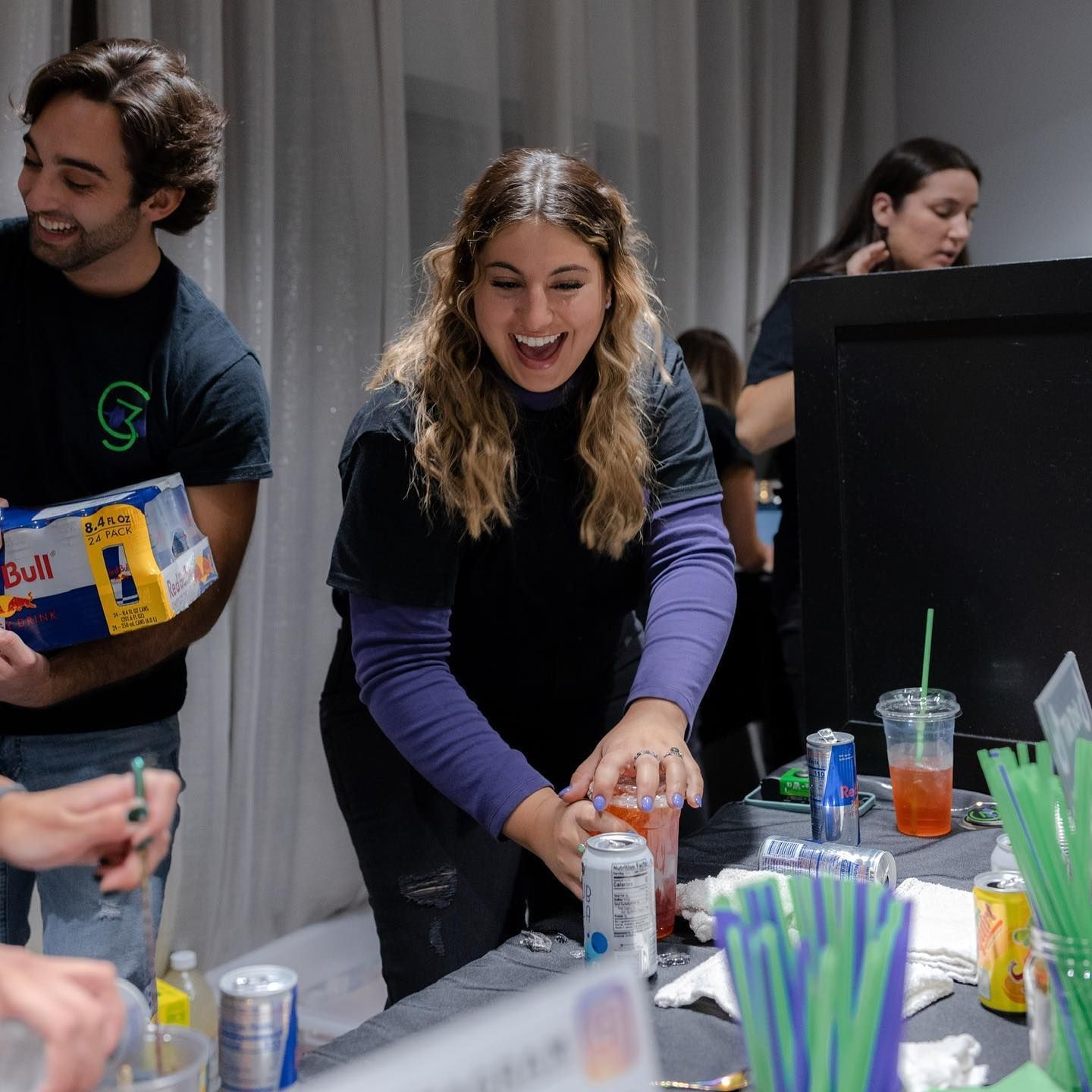 People at a bar, preparing drinks. Woman smiling, mixing a drink, surrounded by cans and straws. Man holding Red Bull.