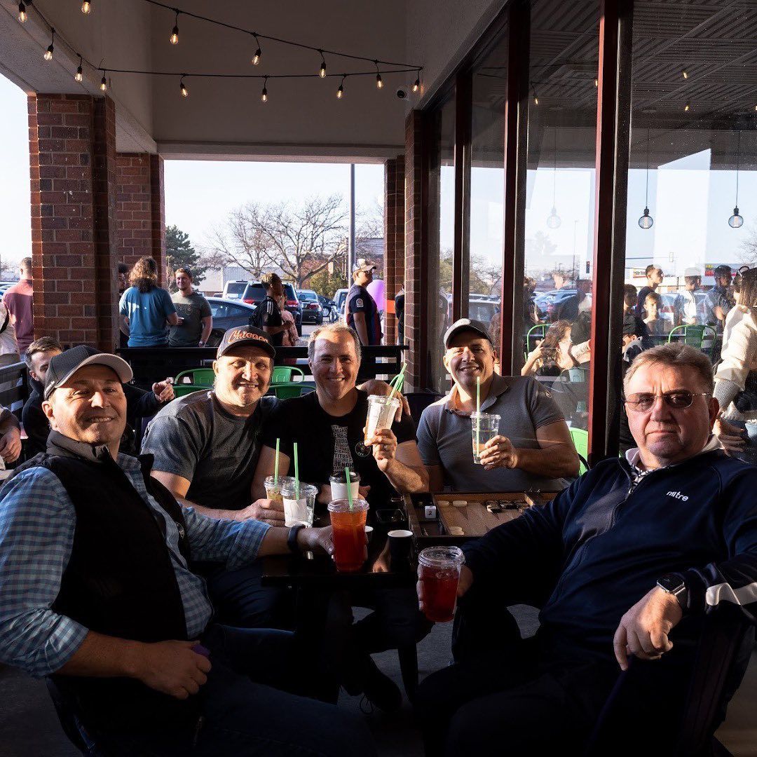 A group of six men outdoors at a table, smiling and holding drinks. Sunlight streams in from behind.