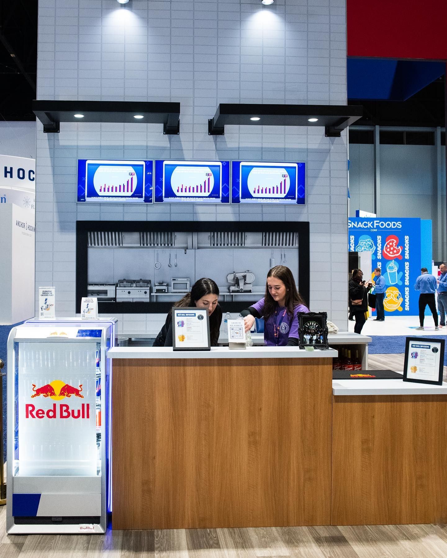 Two people at a wooden counter. Above, three screens show graphs. Red Bull cooler on the left.