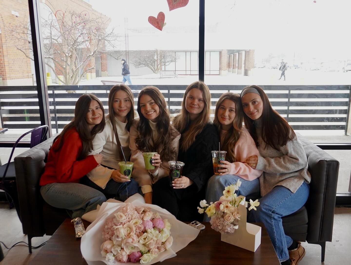 Six people smile, holding drinks, seated on a couch near a window. Flowers and a table are in front of them.