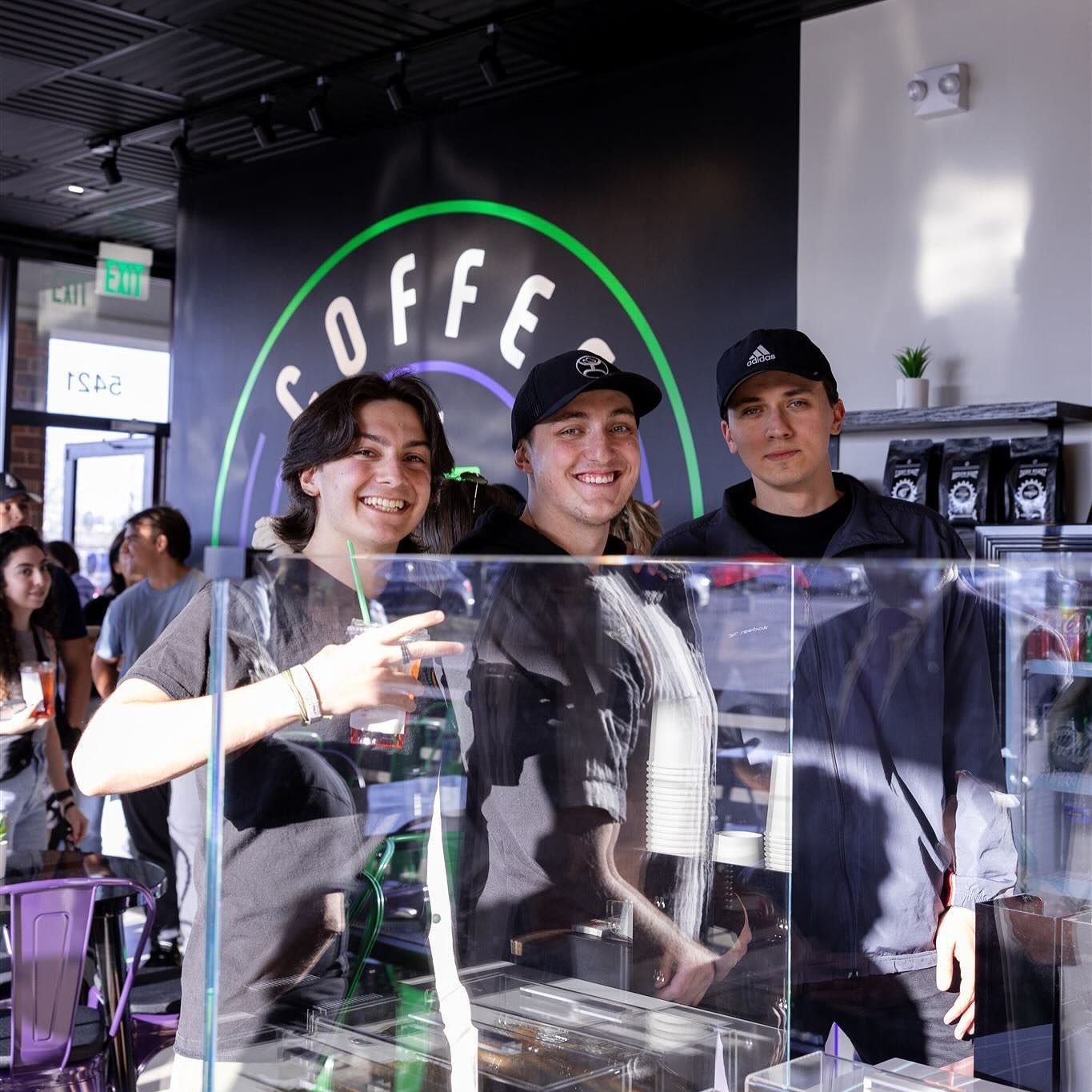Three people behind a coffee shop counter smiling. Coffee shop sign in background.