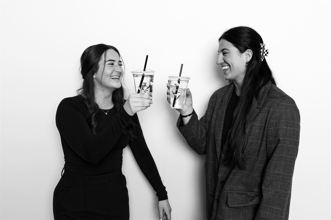Two people toasting drinks, smiling, against a white backdrop.