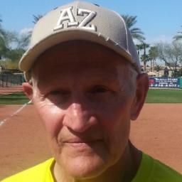 Football coach with arms crossed, looking stern on a sunny field, players in the background.