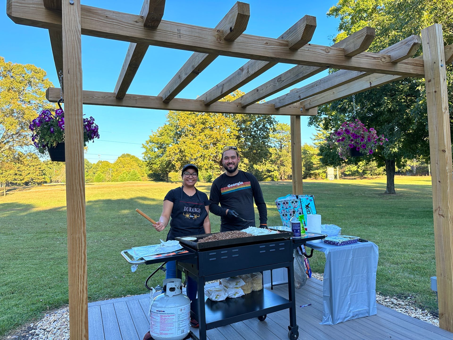 A man and a woman are standing in front of a grill under a pergola.