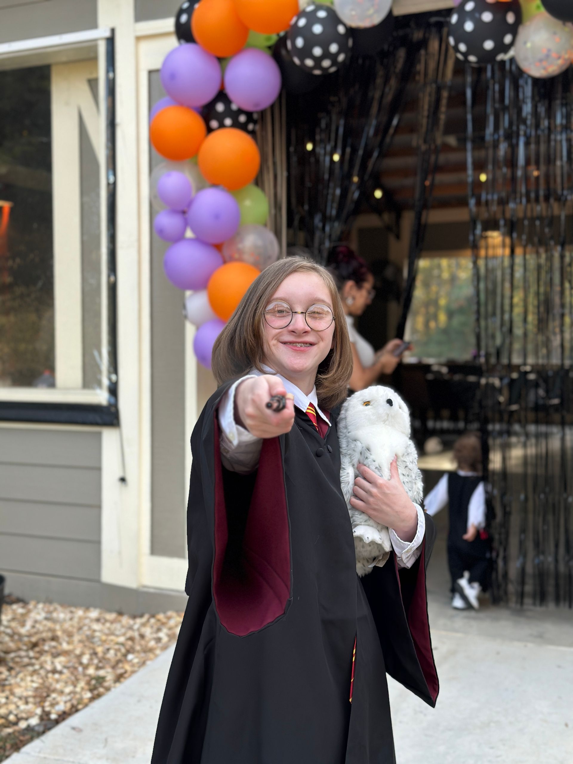 A girl in a harry potter costume is holding a stuffed owl and pointing at the camera.