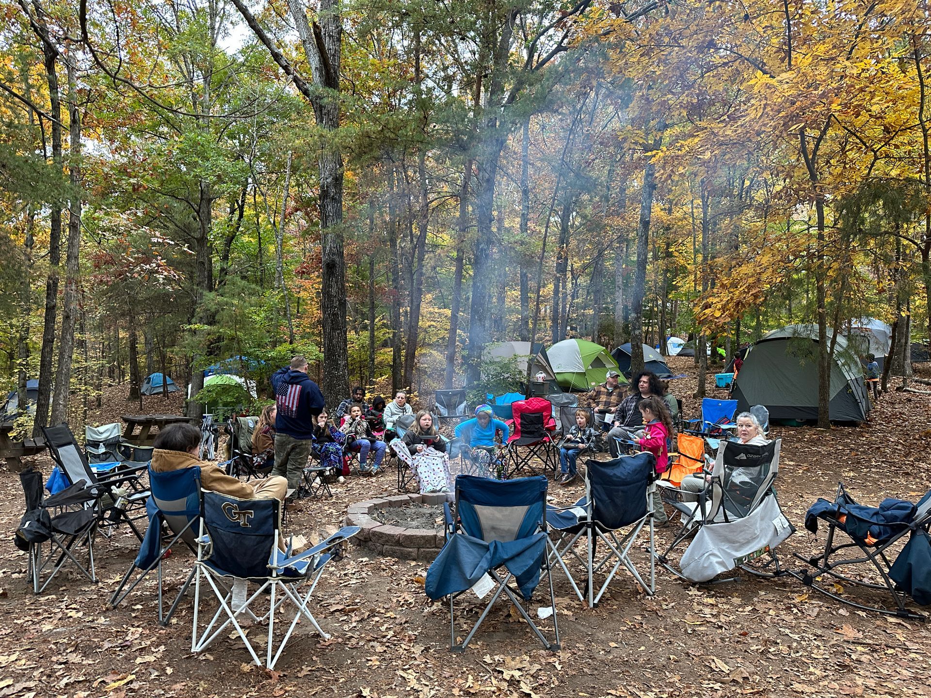 A group of people are sitting around a fire pit in the woods for cub scouts