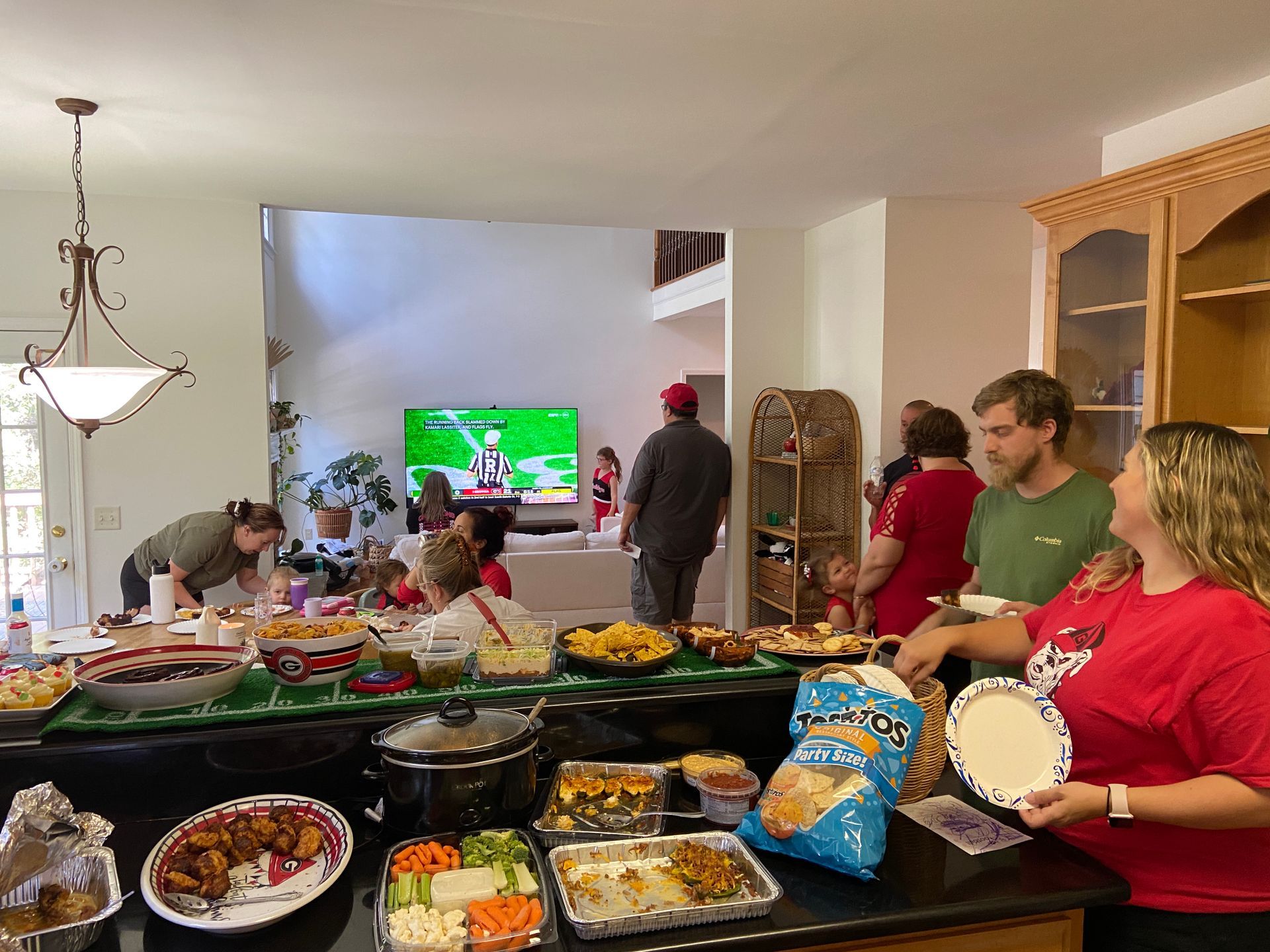 A group of people are standing around a table filled with food watching a football game