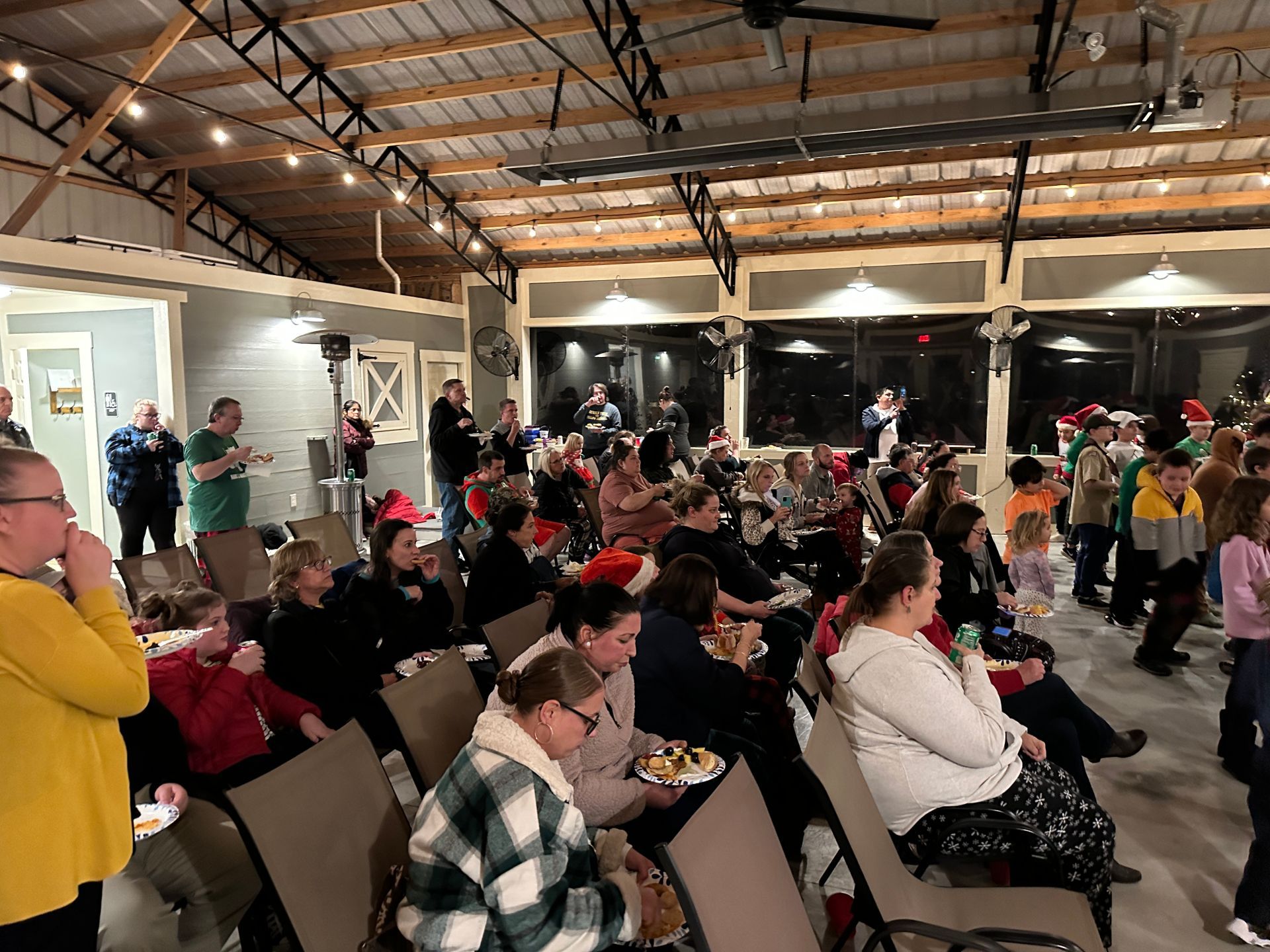 A large group of people are sitting and eating for a Holiday party for cub scouts in a pavilion.