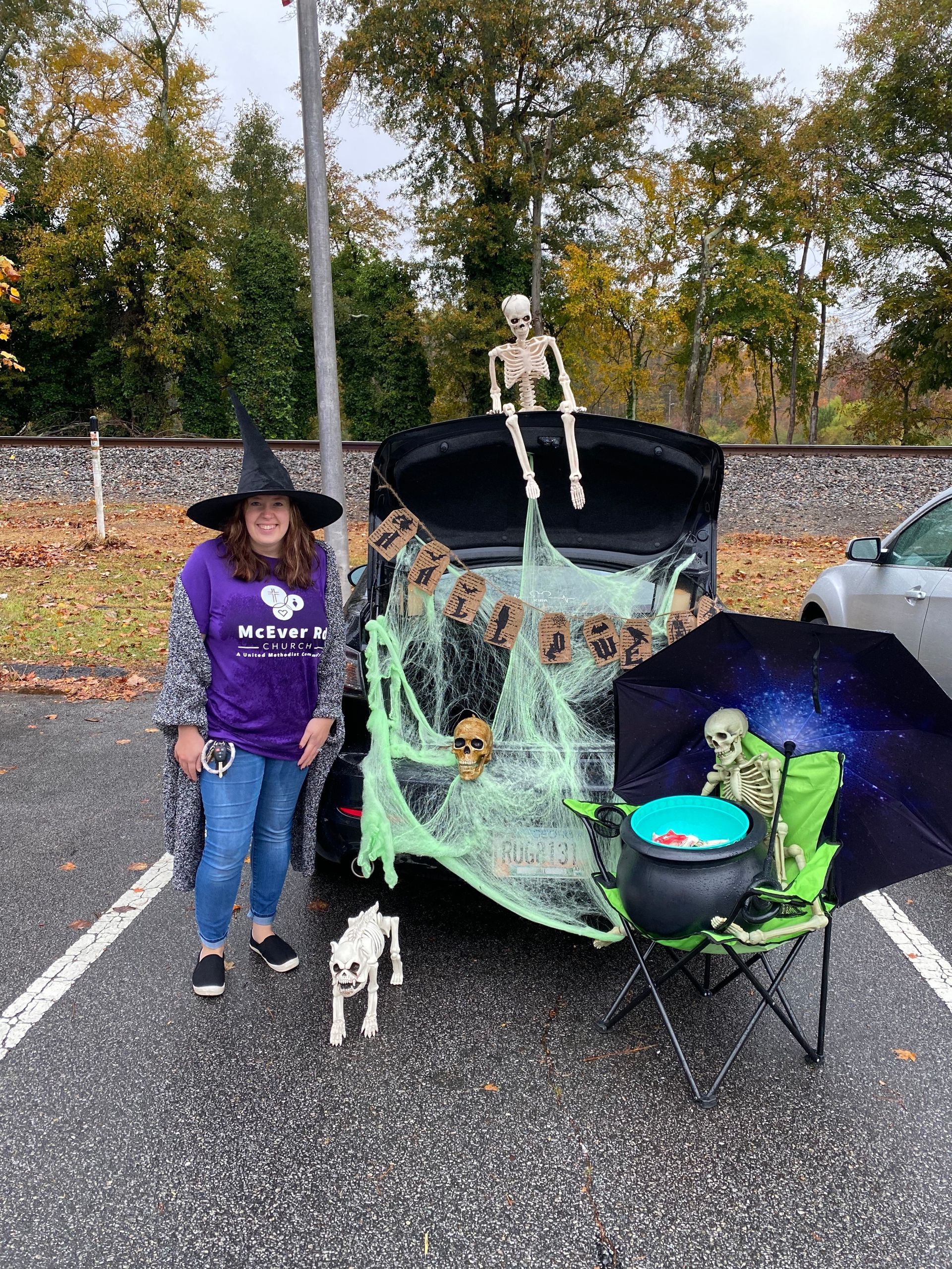 A woman in a witch hat is standing next to a car with a trunk decorated for halloween.