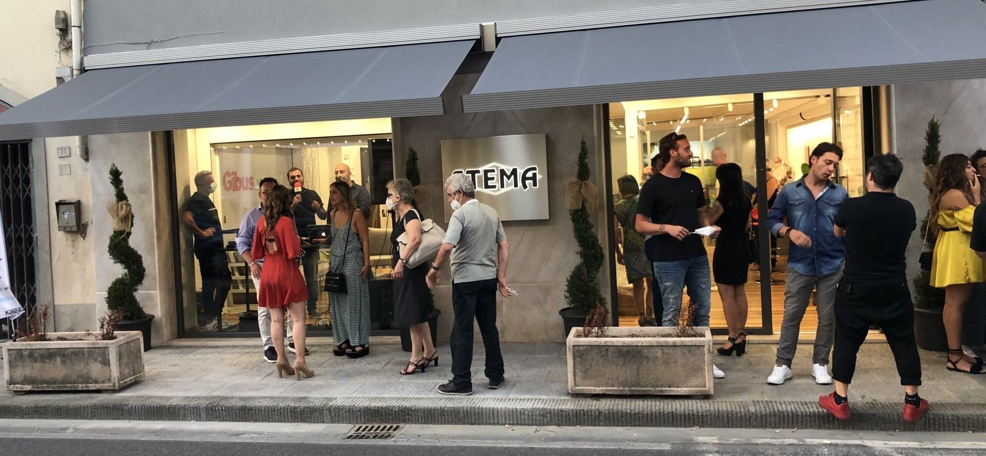Persone radunate fuori da un negozio con una tenda da sole. Un cartello recita 