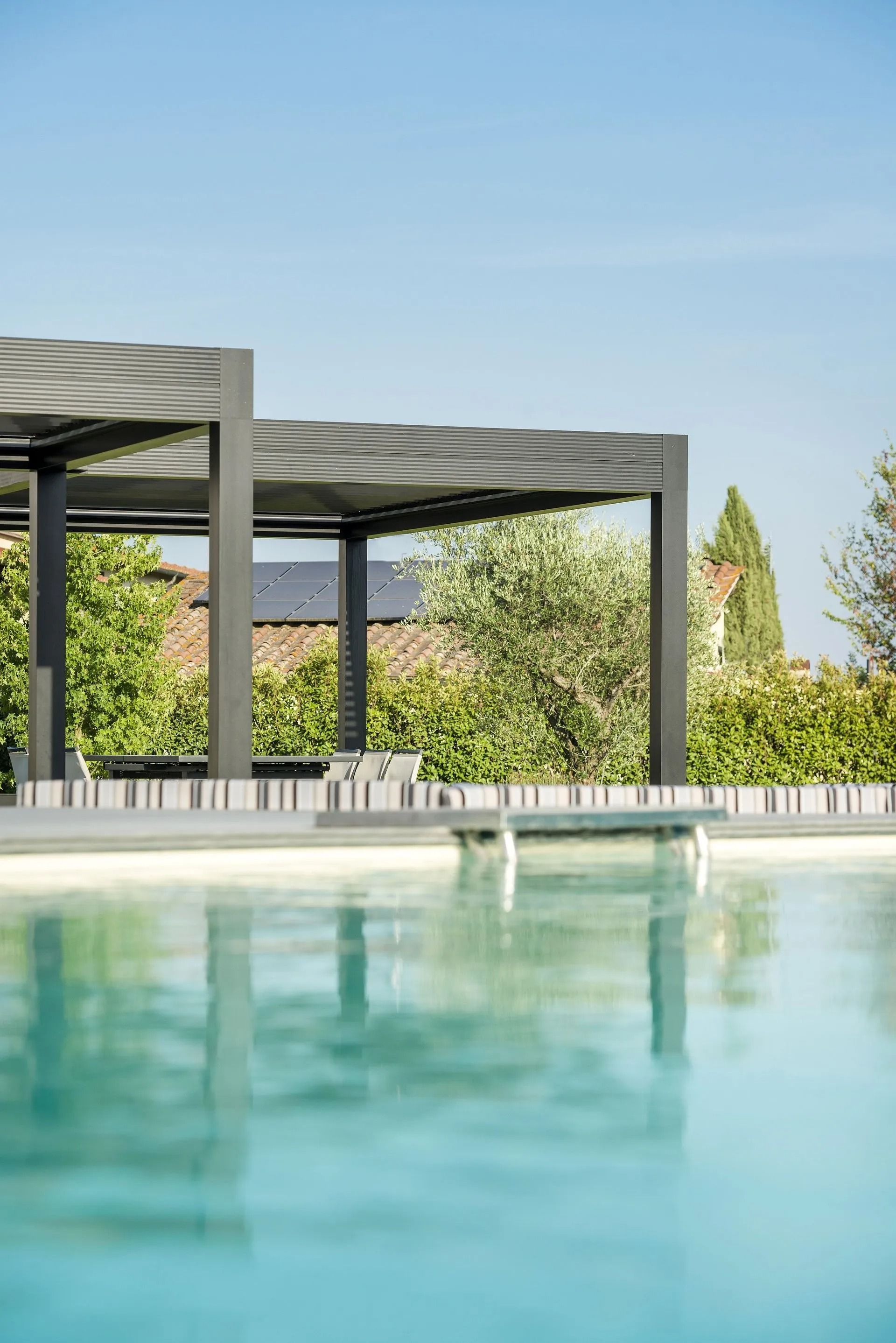 Vista a bordo piscina con superficie riflettente, un pergolato moderno e vegetazione sotto un cielo azzurro.