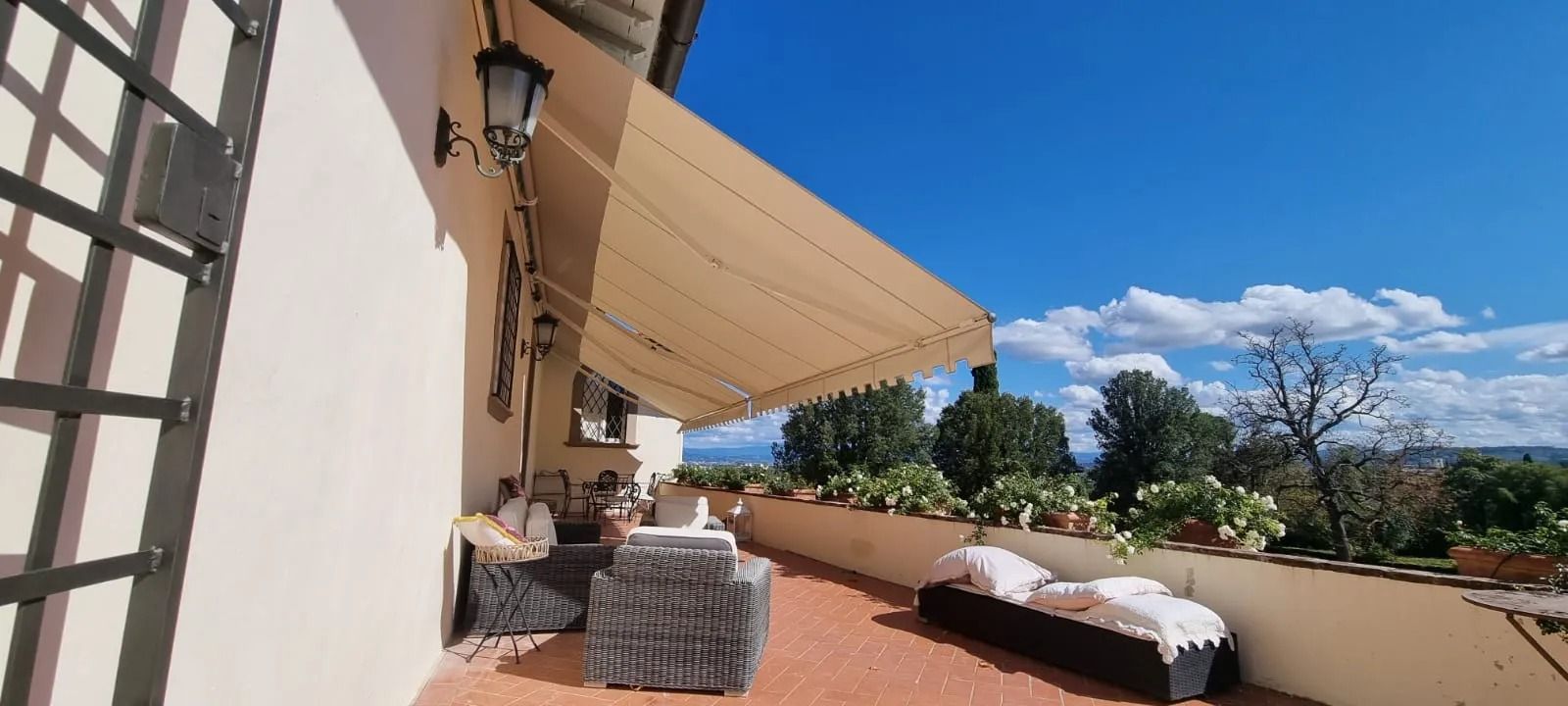 Terrazza con tenda da sole beige, mobili da esterno e fioriere; cielo azzurro sullo sfondo.