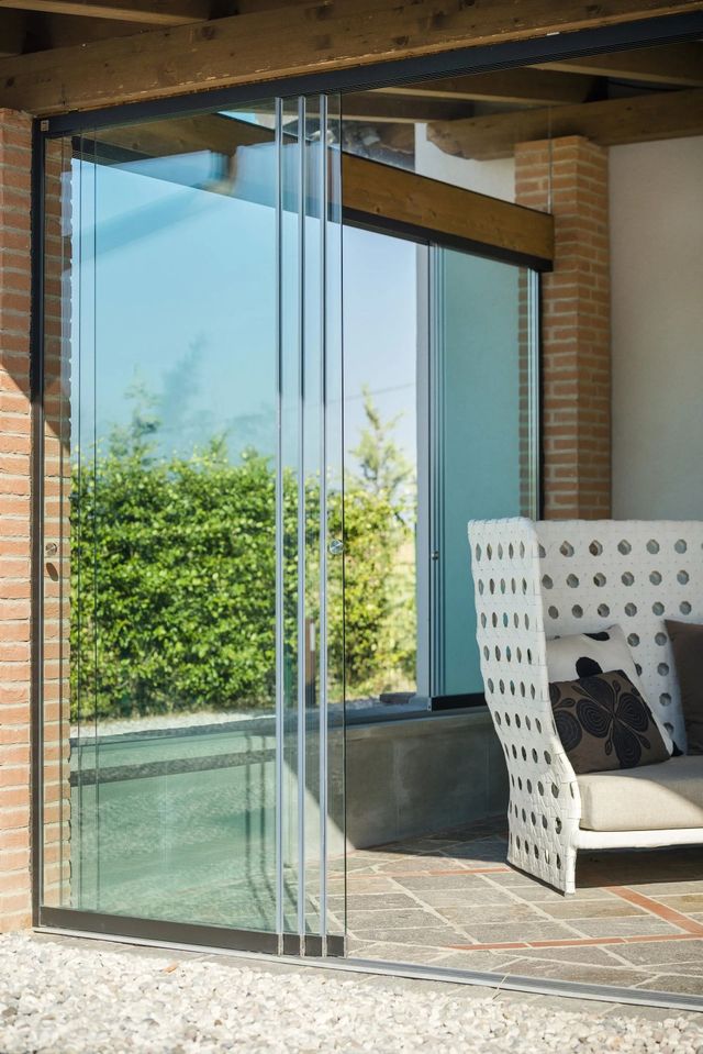 Le porte pieghevoli in vetro si aprono su uno spazio esterno con vegetazione lussureggiante e un divano bianco.