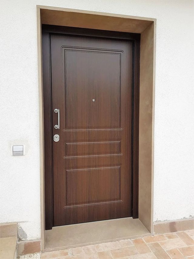 Porta marrone con pannelli decorativi inseriti in una cornice marrone chiaro contro una parete in stucco bianco.