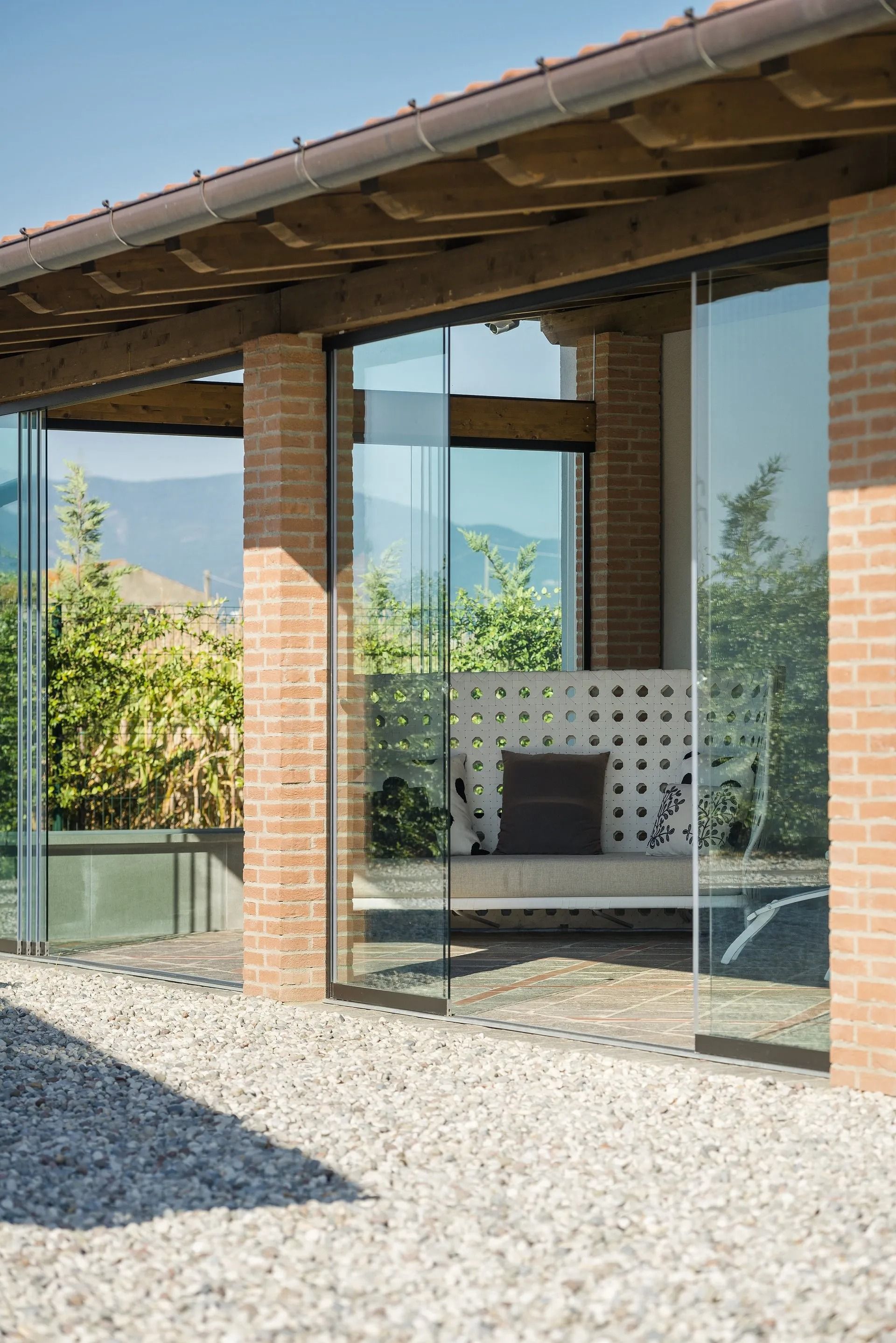 Esterno con pareti in vetro e pilastri in mattoni, che rivelano una zona salotto e un giardino, sotto un tetto