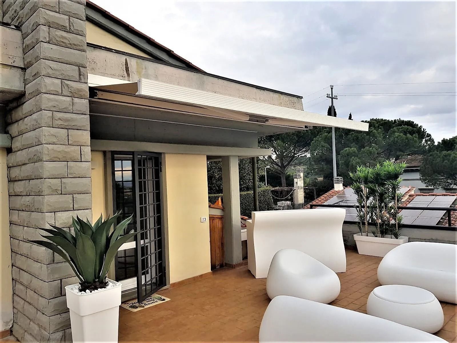 Patio sul tetto con mobili bianchi, piante in vaso e tenda da sole. Pareti in mattoni beige e grigi. Giornata nuvolosa.