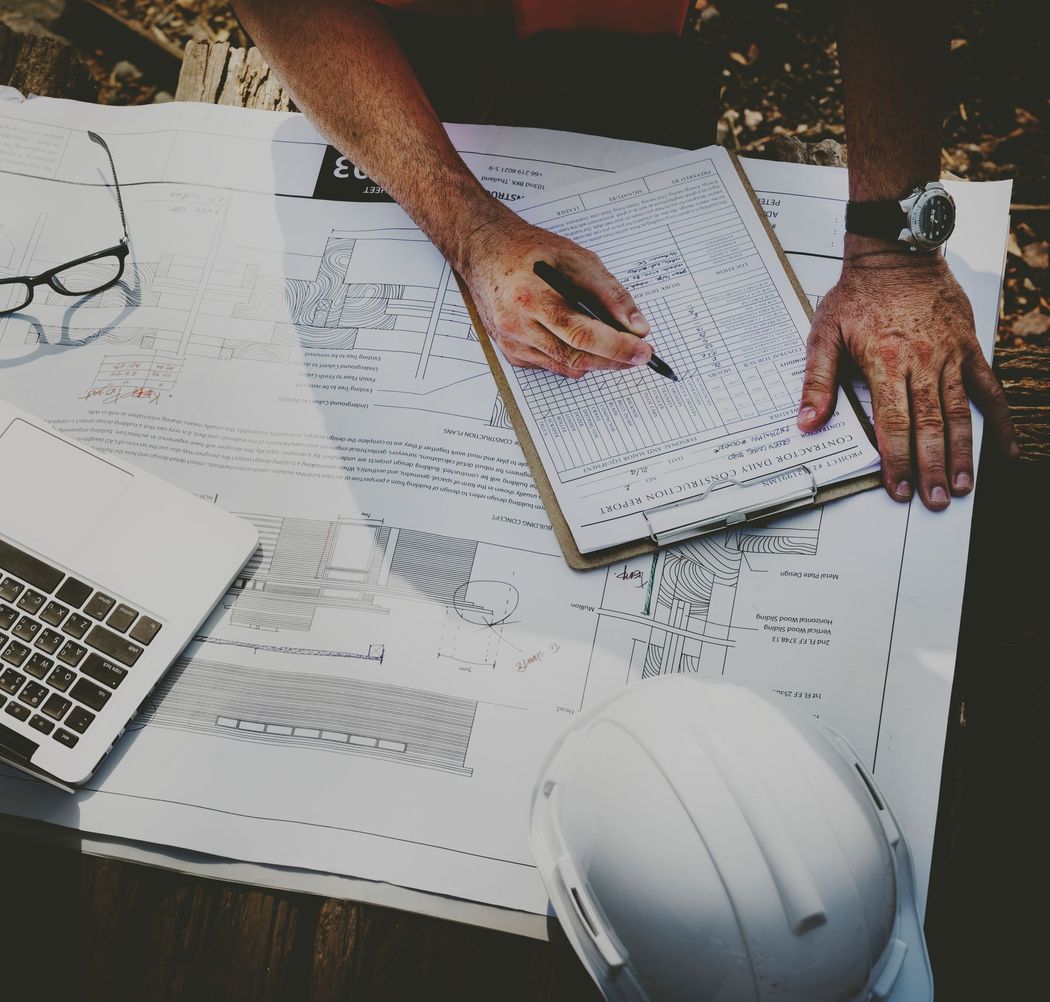 Hands marking plans on a desk with papers, laptop, glasses, and a hard hat