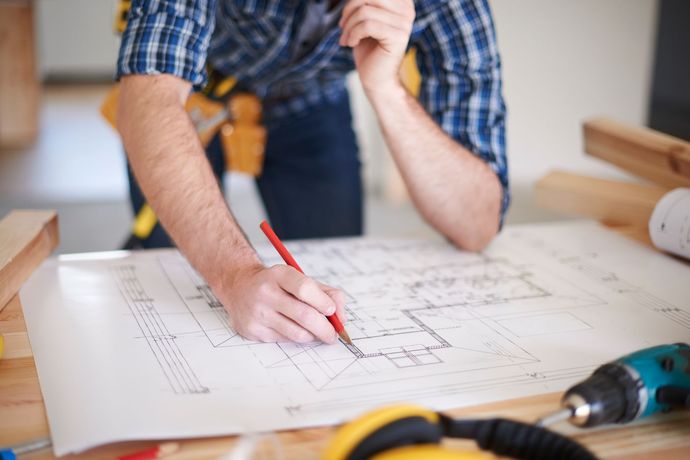 Person drawing on a large architectural blueprint with a red pencil at a workbench