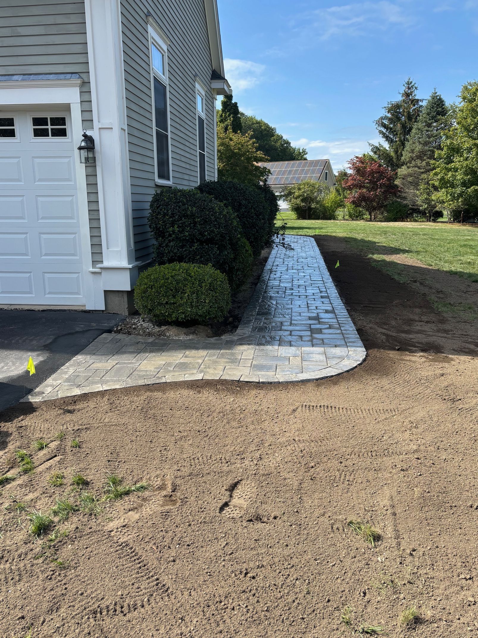 Brick walkway alongside a house with green bushes and a grassy lawn. Blue sky in the background.