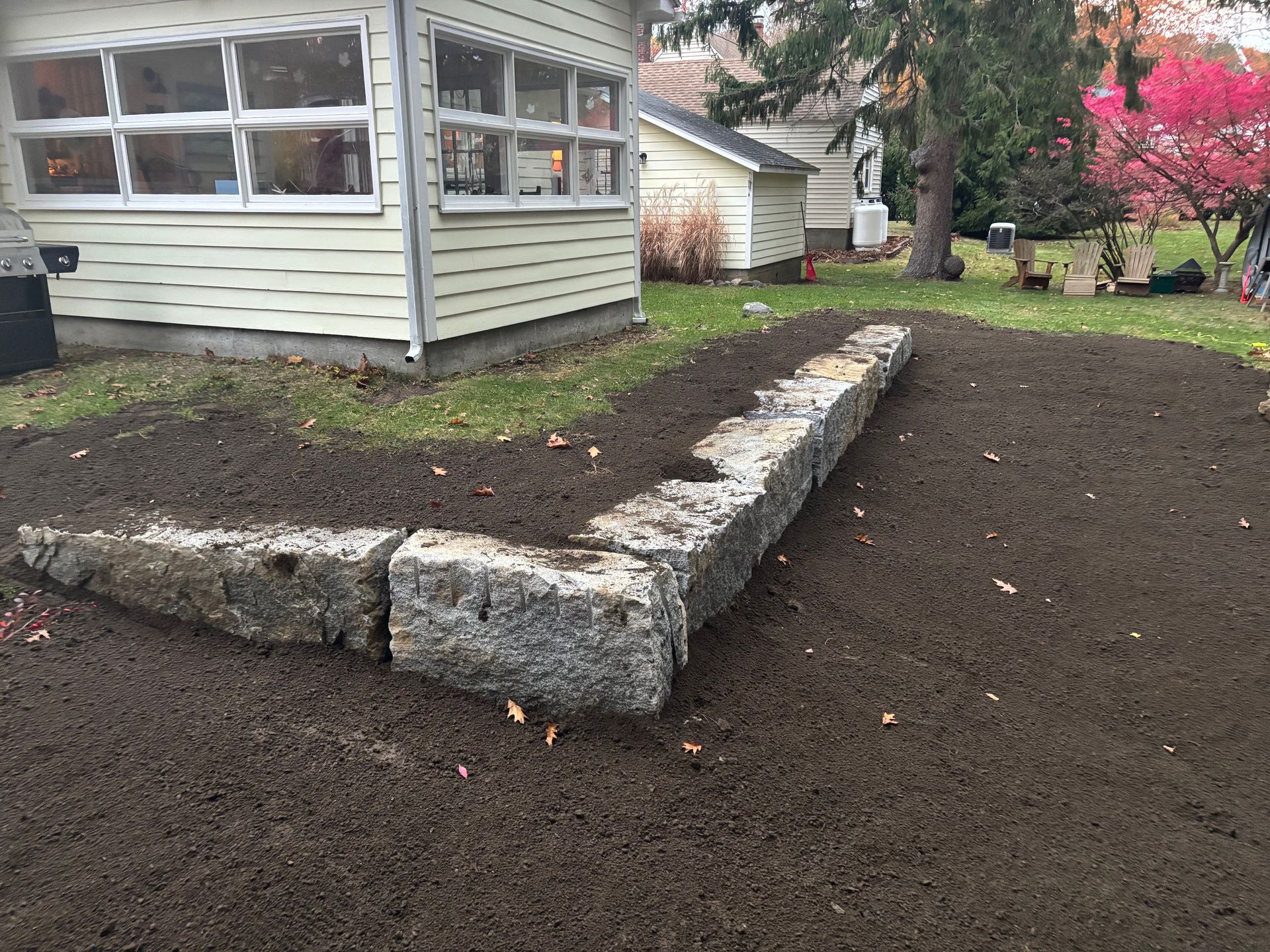 A stone retaining wall in a yard, near a light-colored house. Brown soil surrounds the wall and grass.
