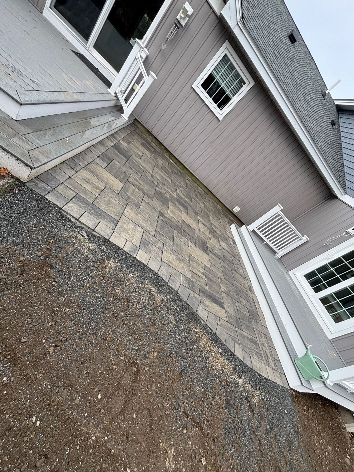 Paver patio beside a light gray house. Gravel path and exposed soil in foreground.