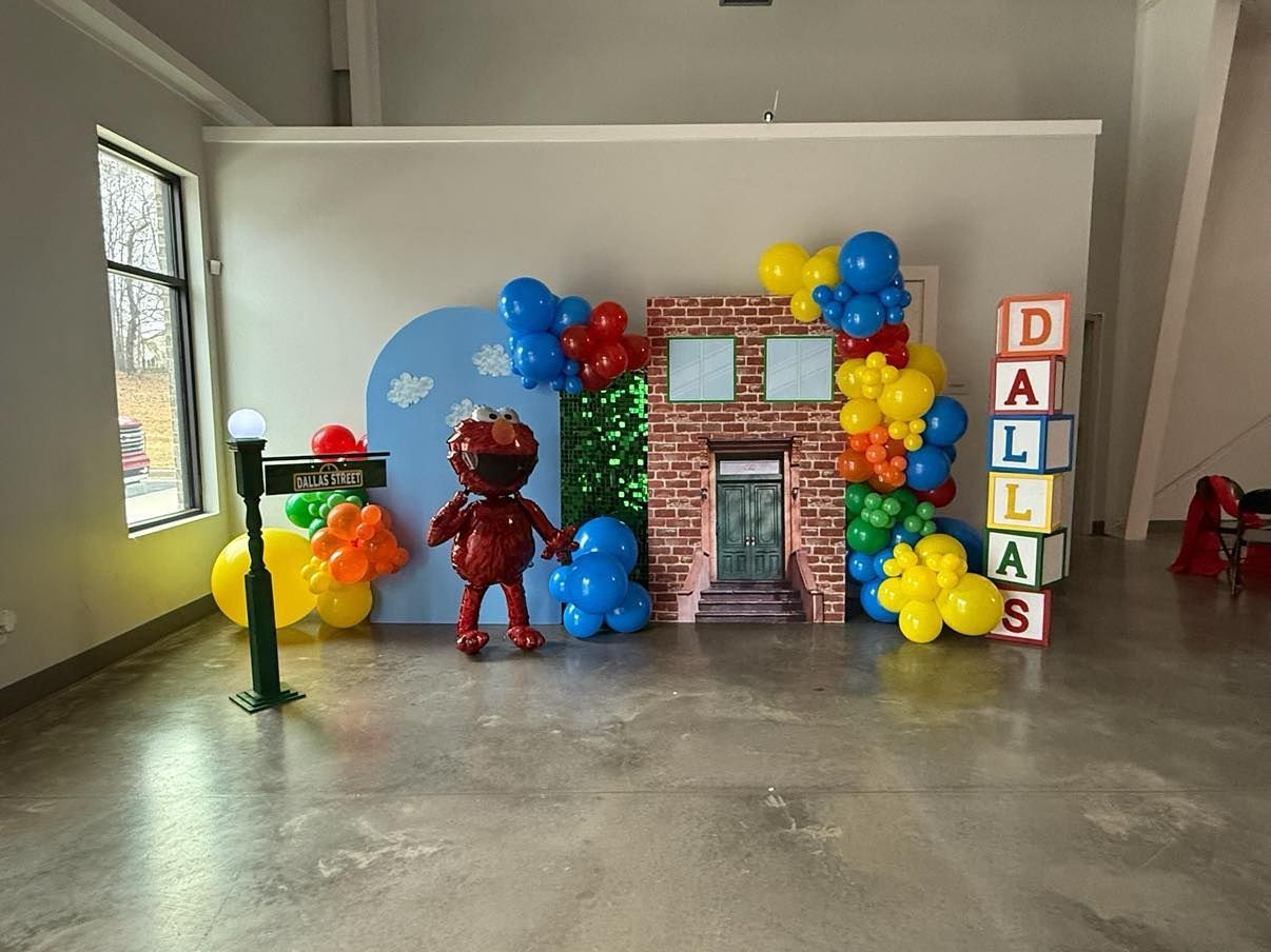 Party backdrop with building, balloons, and a character; blue, red, yellow, and green. 