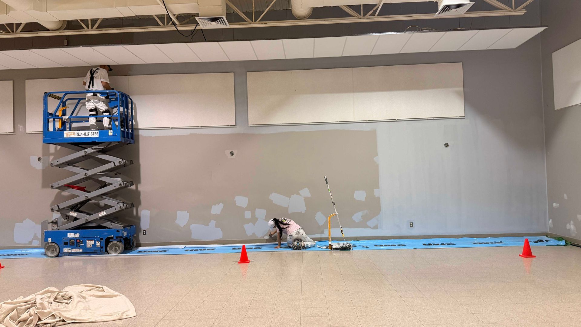Pièce grise vide avec une nacelle élévatrice, du ruban adhésif bleu au sol et des cônes orange devant un mur rafistolé.