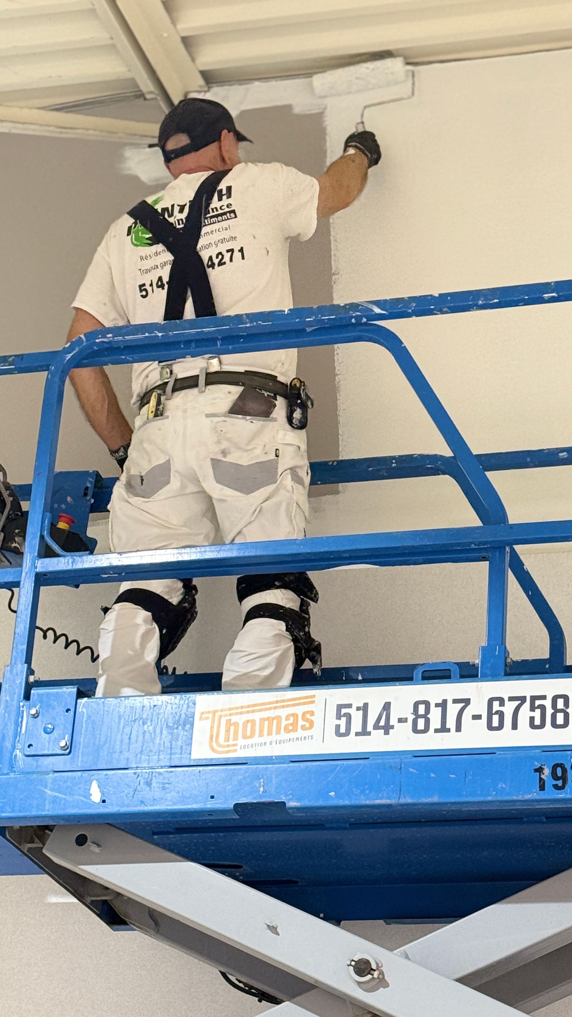 Un ouvrier, perché sur une nacelle élévatrice bleue, peint un plafond blanc à l'intérieur.