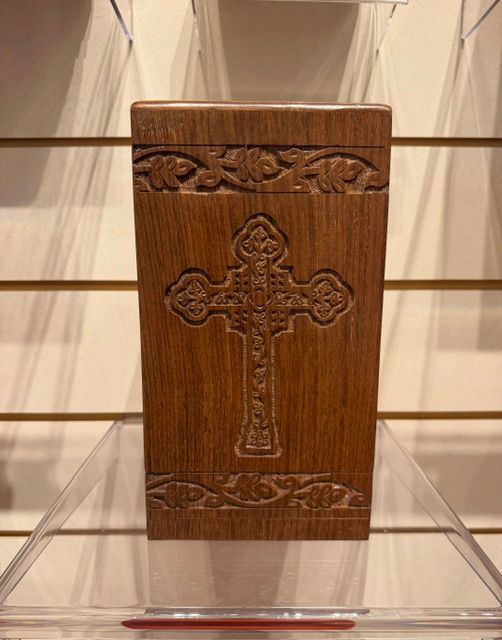 Wooden religious box with an engraved cross on a clear display stand, against a slatwall background