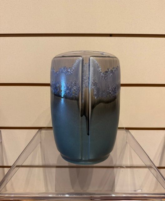Blue-gray glass vase with a dark vertical stripe, displayed on a clear stand against a slatwall background