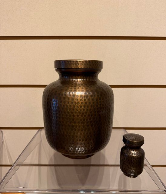 Two dark bronze patterned jars on a clear acrylic stand against a beige slat wall.