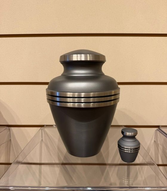 Two black urns with silver bands displayed on a glass shelf against a beige wall.