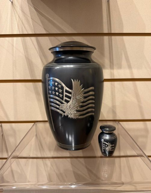 Black urn with silver eagle and flag design, beside a small matching mini urn on a shelf.