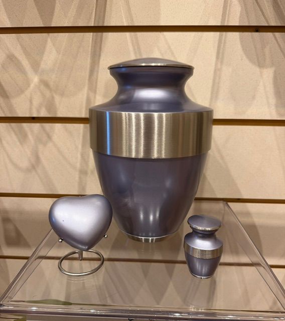 Three silver funeral urns, including a large central urn, displayed on a glass shelf.