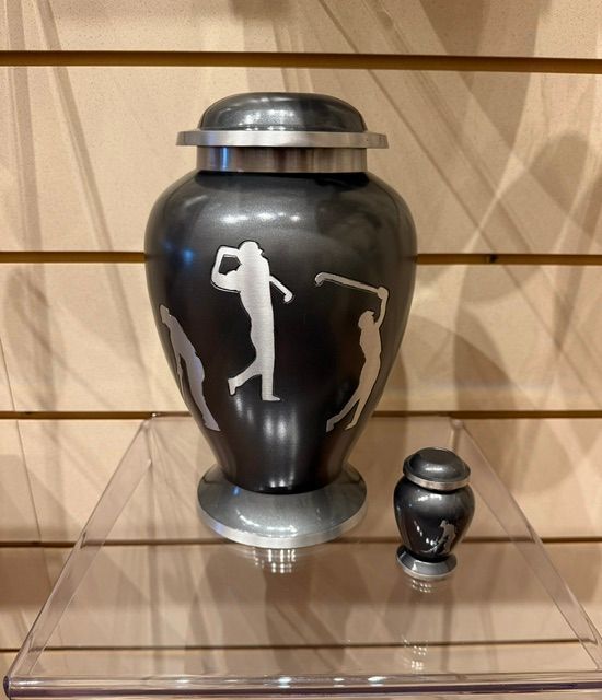 Black urn with silver lid and base, decorated with golf silhouettes, on a glass shelf beside a small matching urn.
