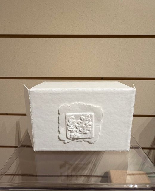 White decorative box with a raised square emblem, displayed on a clear stand against a slatted wall.