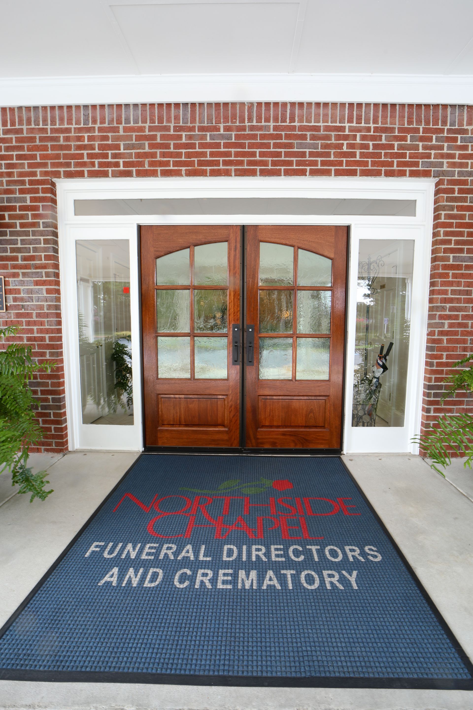 Entrance to a brick funeral home with wooden double doors and a black mat reading “Jenkins Funeral Directors and Crematory”