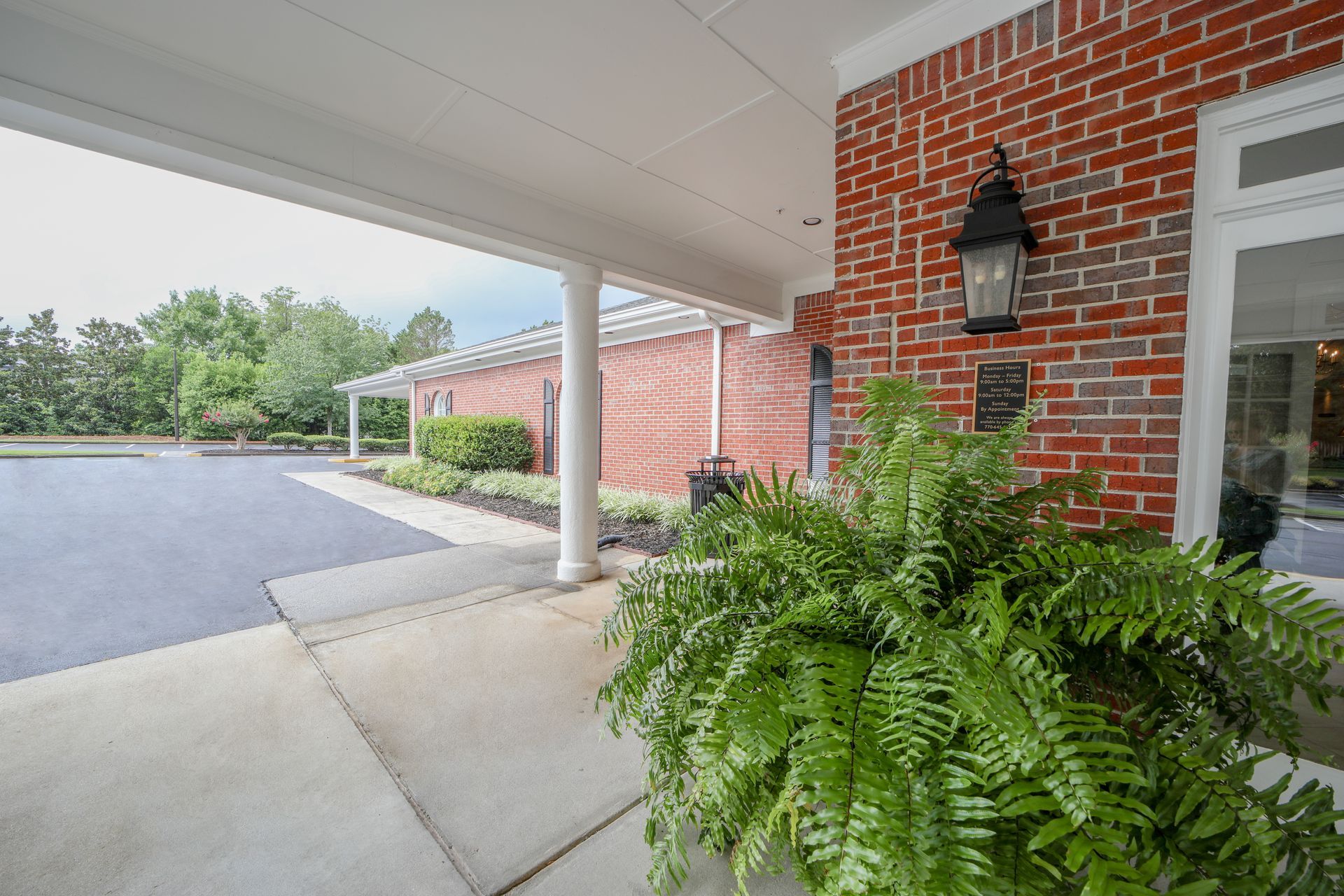 Covered brick entryway with white columns, fern planter, and driveway beside a building