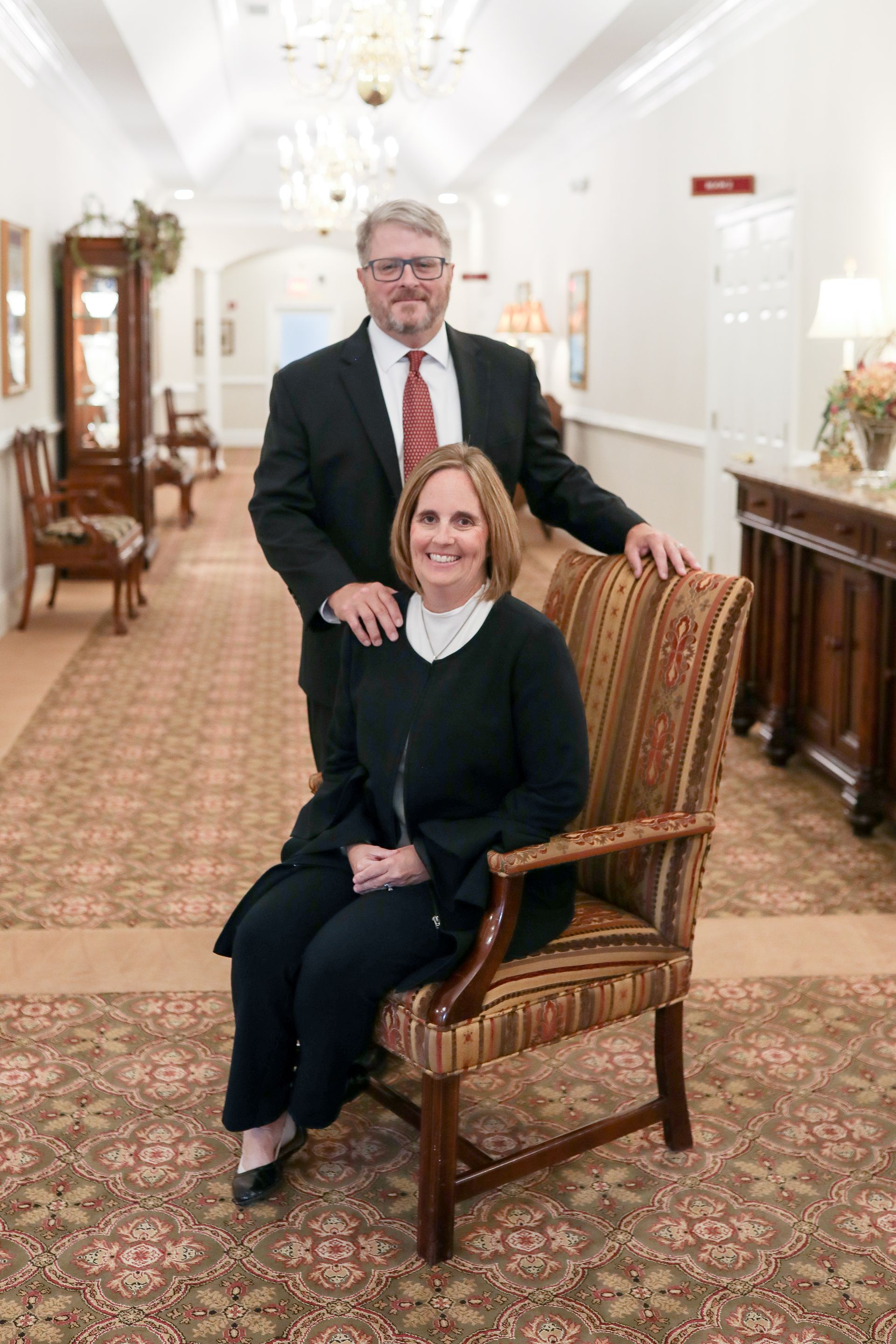 Two people posing in a formal hallway, one seated in a patterned chair and one standing behind, both smiling.