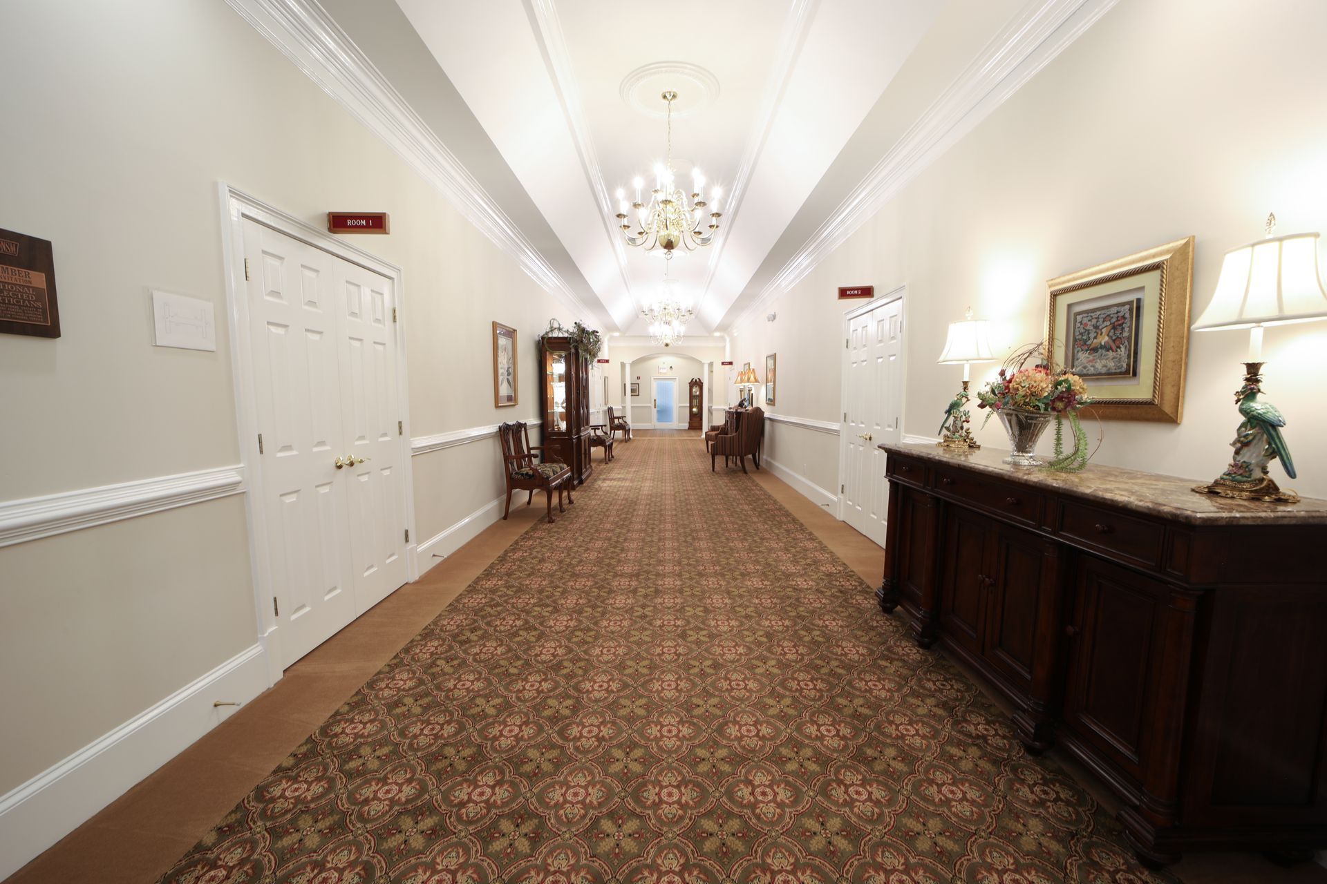 Elegant hotel hallway with carpet, chandeliers, and dark wood console tables along white walls