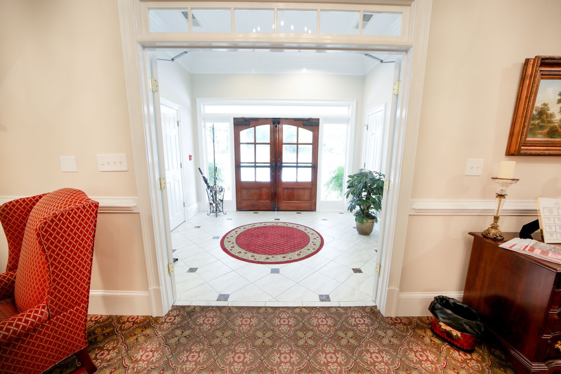 Bright foyer with double wooden doors, red oval rug, plants, and patterned carpet leading into a white hallway