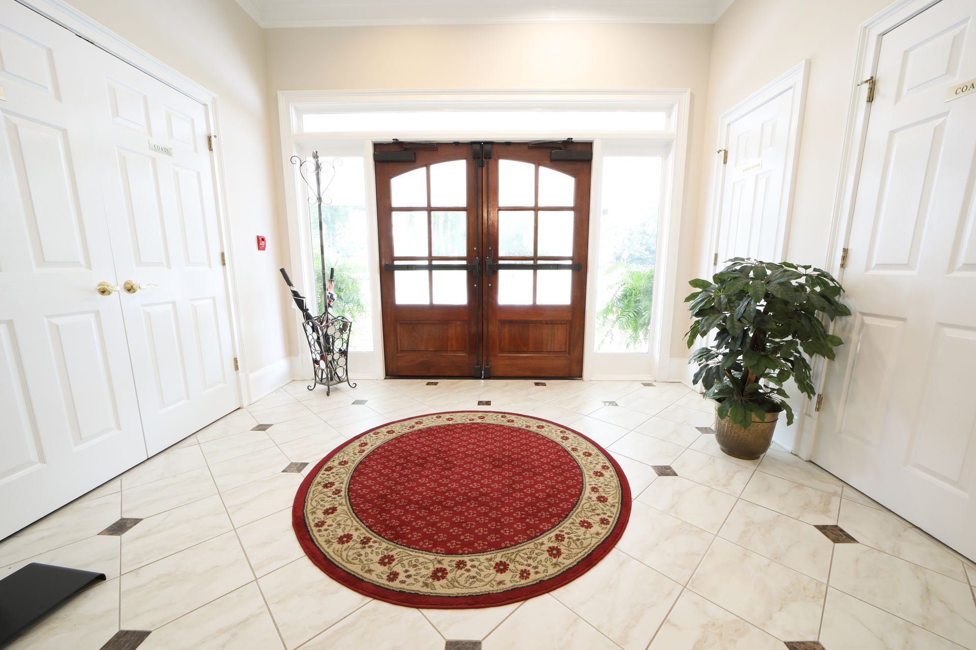 Bright foyer with double wooden doors, white walls, marble floor, red round rug, and potted plants.