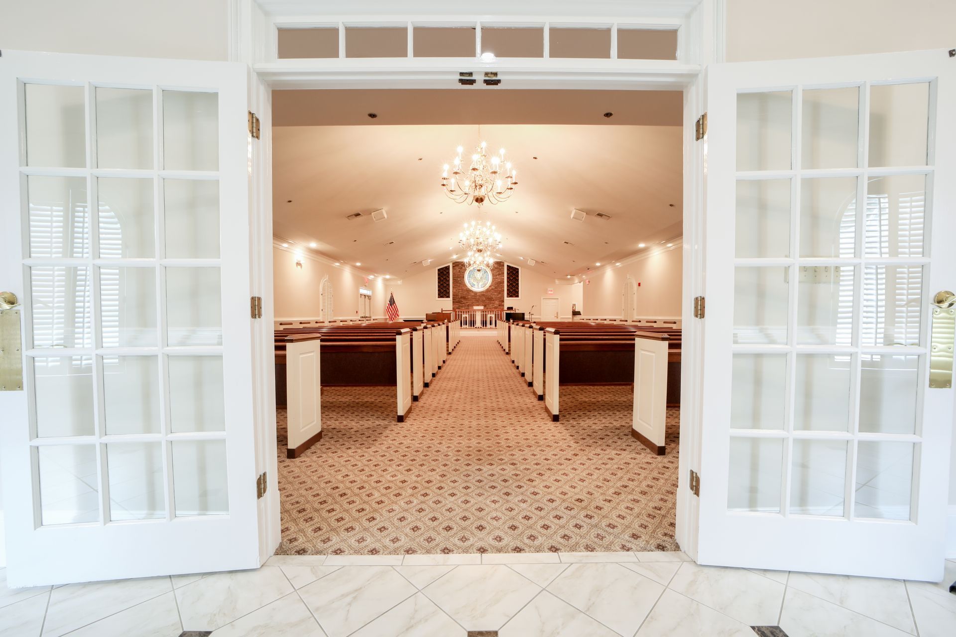 Open double doors to a bright, elegant hall with chandeliers and rows of chairs.