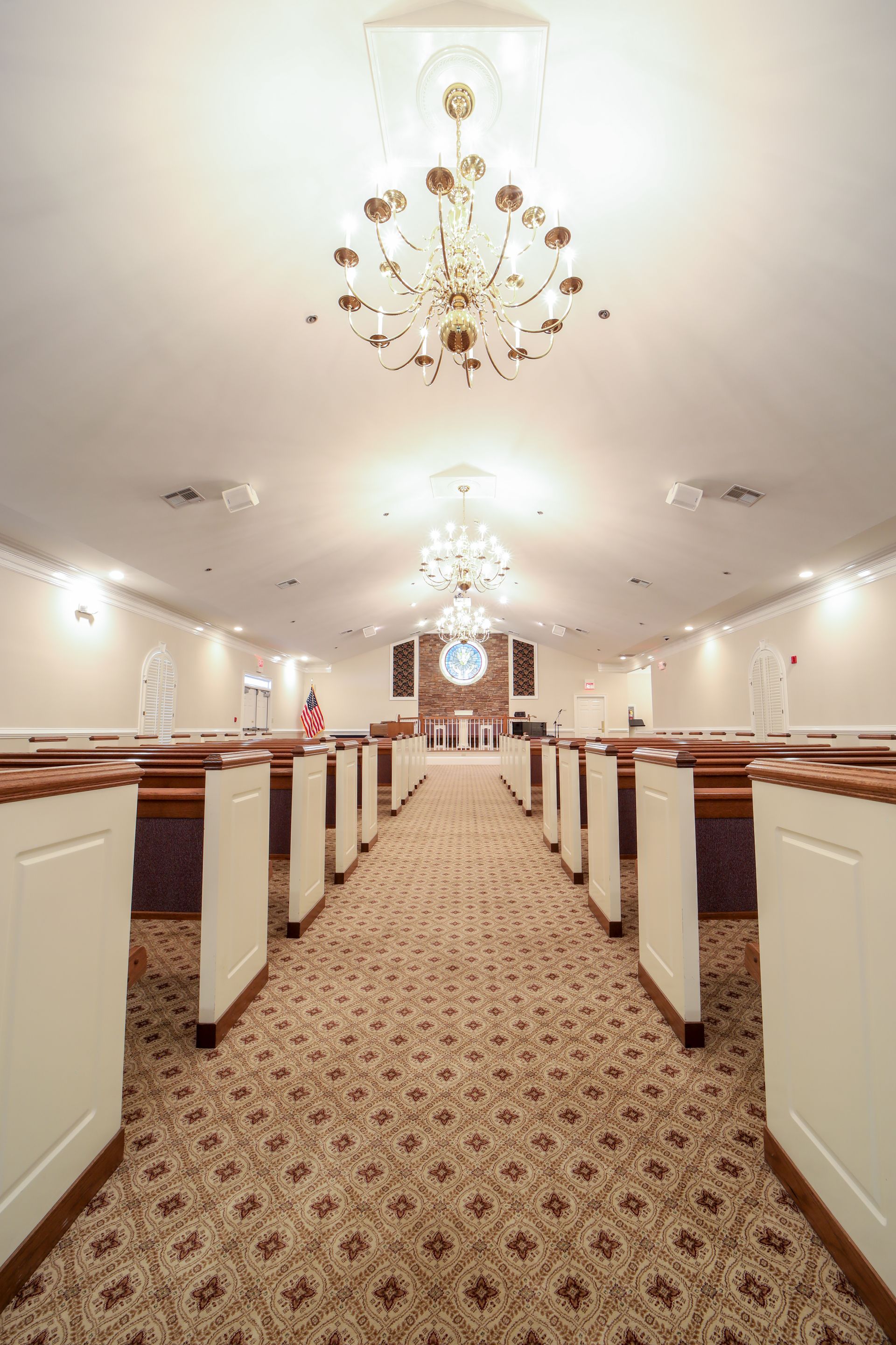 A church aisle with patterned carpet, white pews, and chandeliers under a bright ceiling.