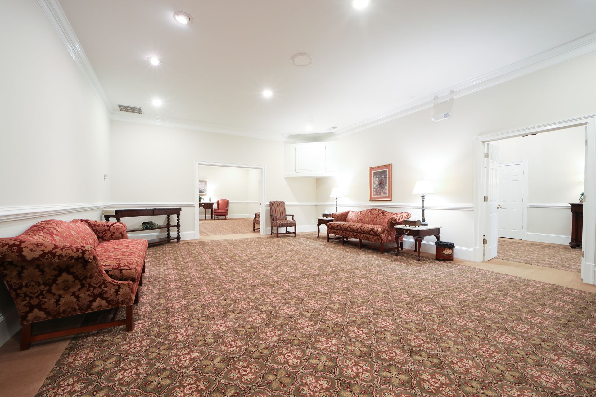 Spacious white living room with patterned carpet, red sofas, tables, and recessed ceiling lights