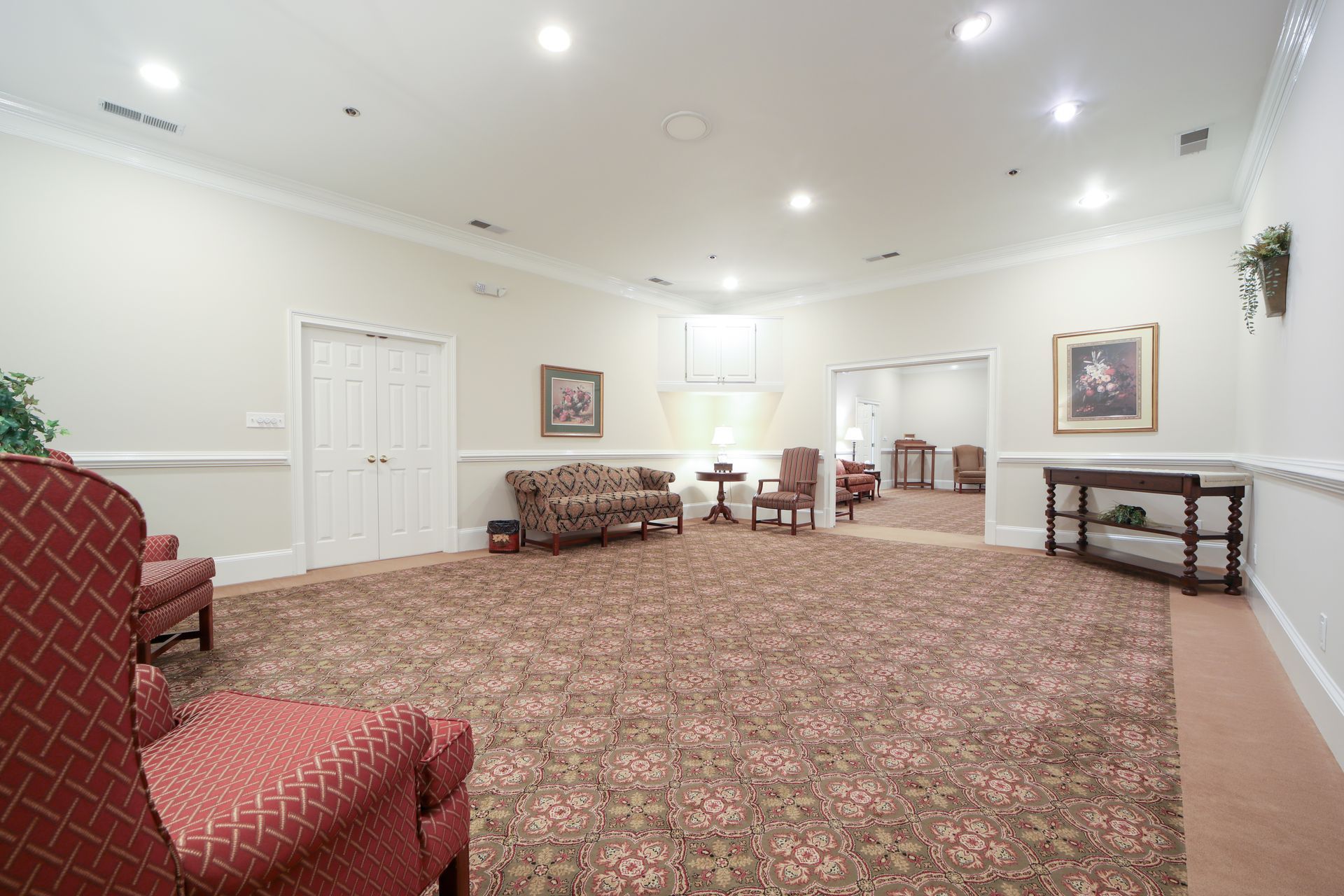 Spacious carpeted lobby with chairs, benches, tables, and framed wall art under bright ceiling lights