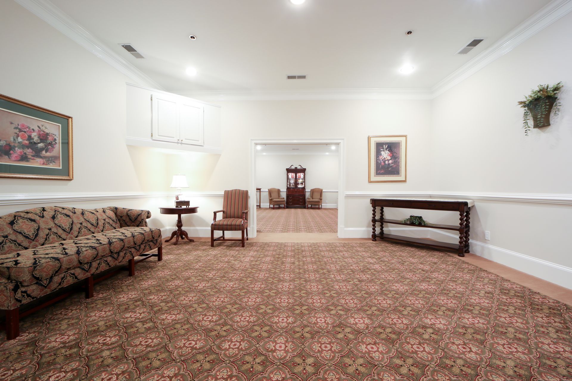 Empty carpeted lobby with white walls, framed art, a sofa, chairs, and a small table under bright ceiling lights