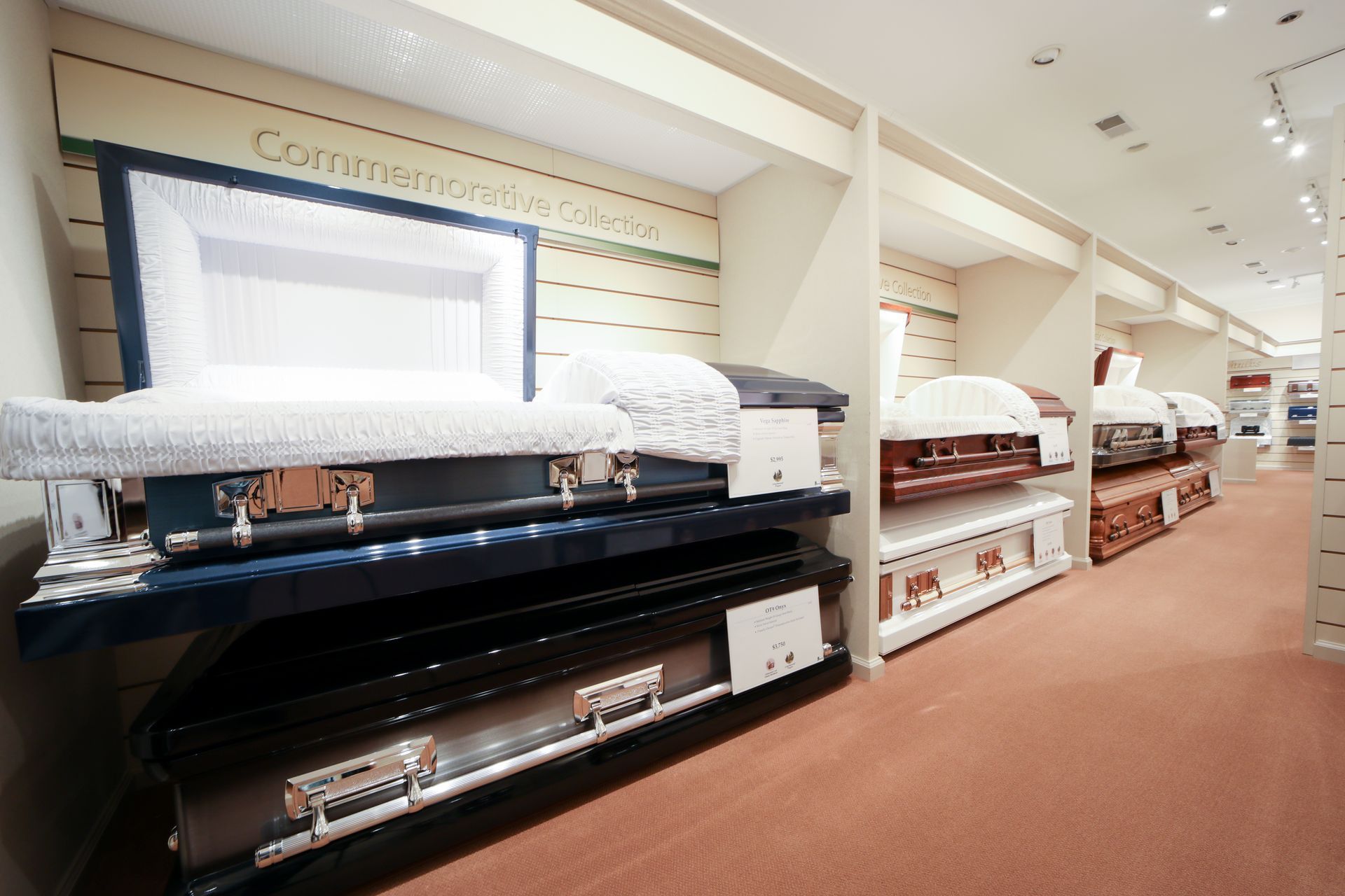 Caskets displayed in a showroom aisle with stacked bedding and shelves under bright store lighting