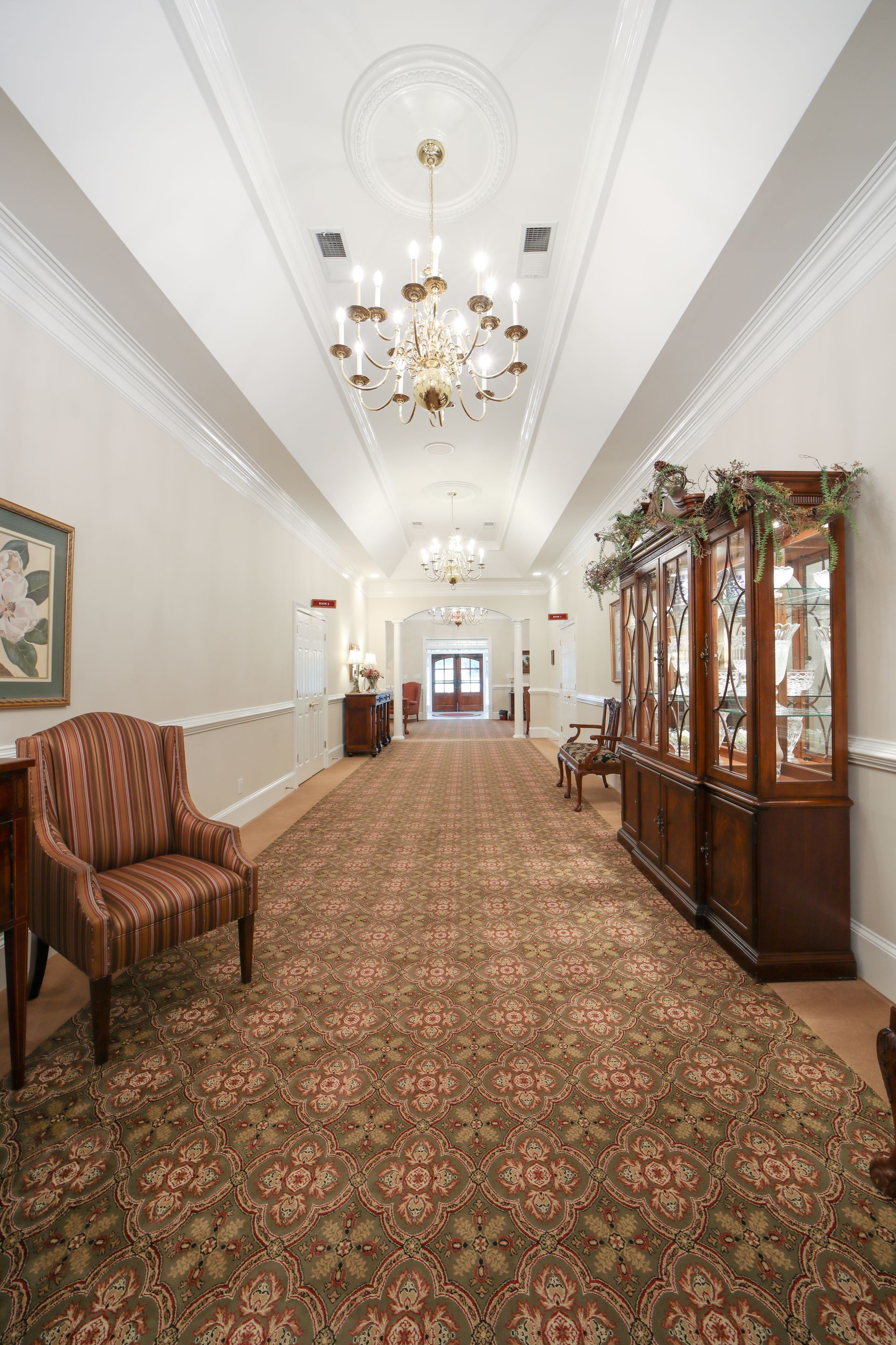 Elegant carpeted hallway with chandeliers, white walls, and antique wooden furniture and cabinets