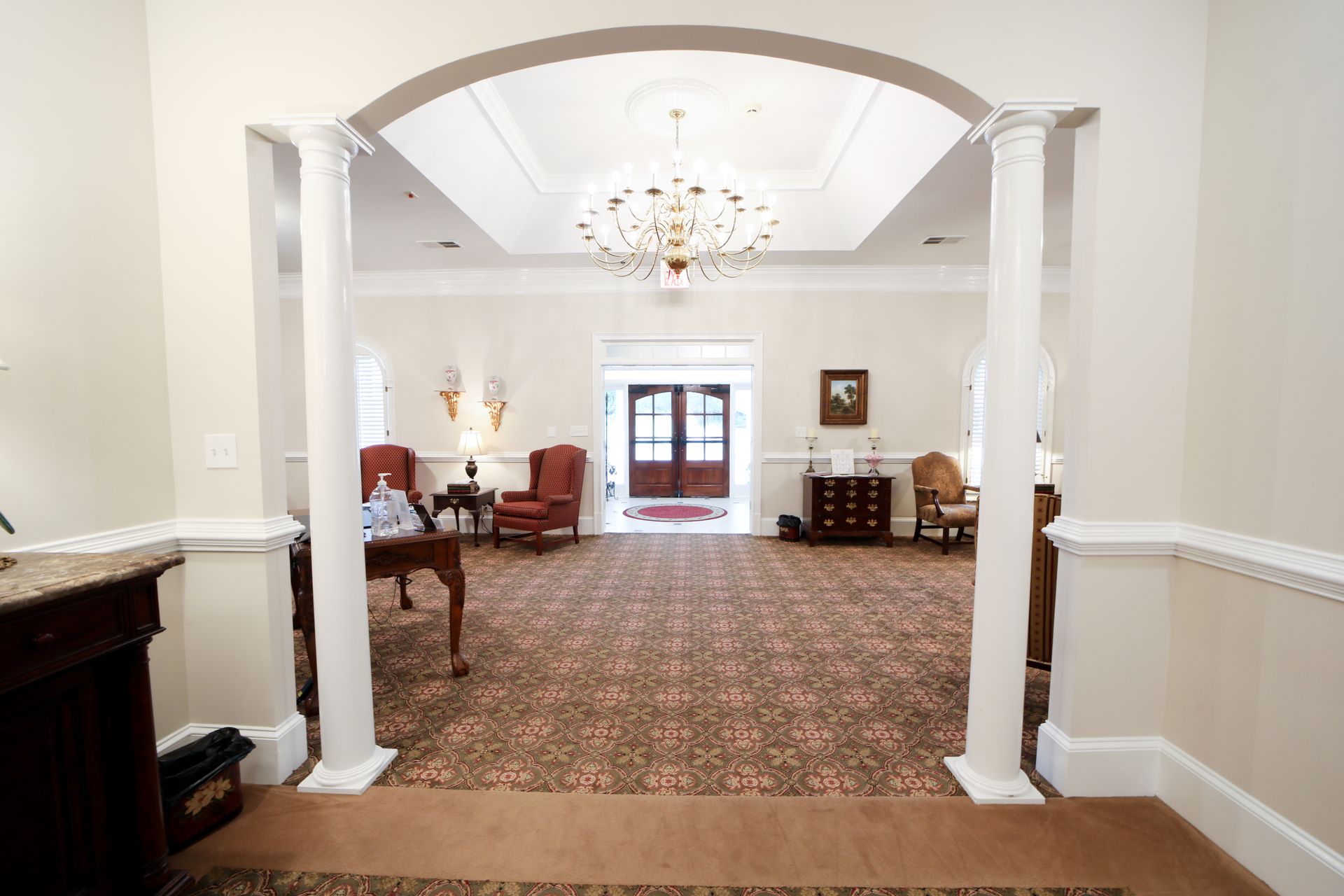 Elegant foyer with chandelier, arched doorway, patterned carpet, and chairs leading to a bright room